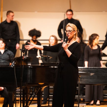 Choir director Dr. Erin Colwitz addressing the audience, choir students and accompanist in background