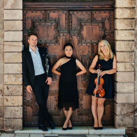 Man in black suit with light blue shirt holds clarinet, pianist stands with arms and legs crossed, and woman with blonde hair holds a violin at her front in front of a large, fancy wooden door.