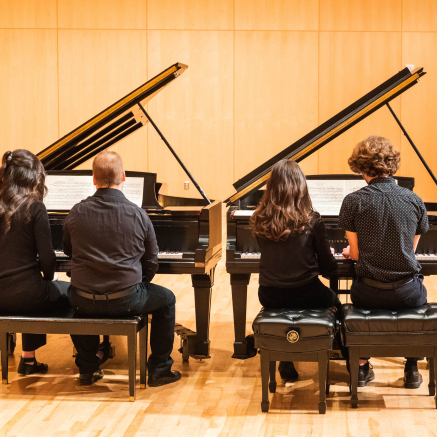 Two pairs of painists sit at two grand pianos, backs facing the viewer